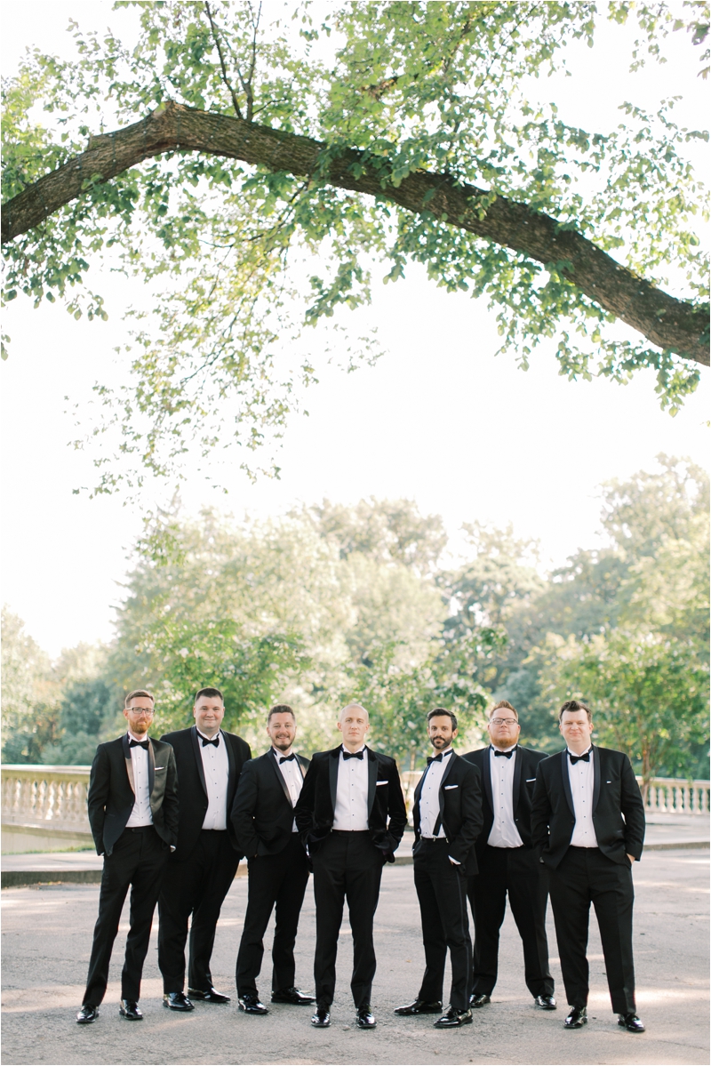 groom poses with groomsmen in tuxes on patio at Curtis Arboretum