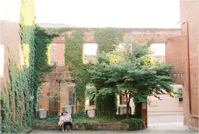 engaged couple sits in front of brick building with green moss